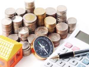 Large stacks of coins with toy house, compass, calculator, and pen.